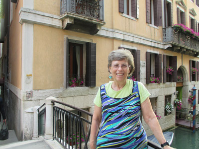 Photo shows Dona standing by the railing of a bridge about 20 feet long, behind her is the corner of a building at least 3 stories tall, with flowers in each window.